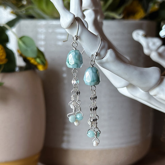 Two earrings hanging from a skeleton hand jewelry stand with tiny dainty faceted Amazonite earrings with pear shaped beads. Each has 3 sterling silver dap chains with small Amazonite and pearl beads dangling on the ends resminiscent of a jellyfish.