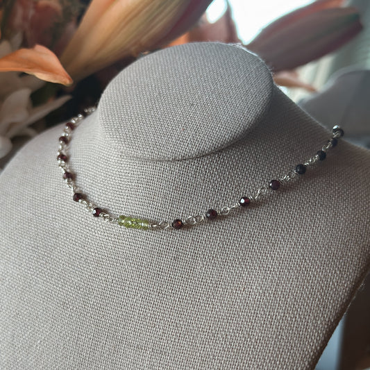 A linen jewelry stand displaying a delicate wire wrapped sterling silver choker featuring high saturation natural Arizona peridot with 5 focal faceted rondelle beads and faceted garnet around the chain.