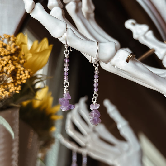 Two sterling silver dangle earrings hanging from skeleton hand jewelry stand each featuring five tiny faceted Amethyst beads in an ombre pattern and a small stack of three unpolished raw Amethyst chips. 